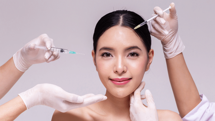 Woman receiving cosmetic injections on her forehead and cheeks from medical professionals. She is smiling and appears relaxed, with gloved hands holding syringes and another hand gently supporting her chin, making one wonder what is Jalupro used for in enhancing natural beauty.