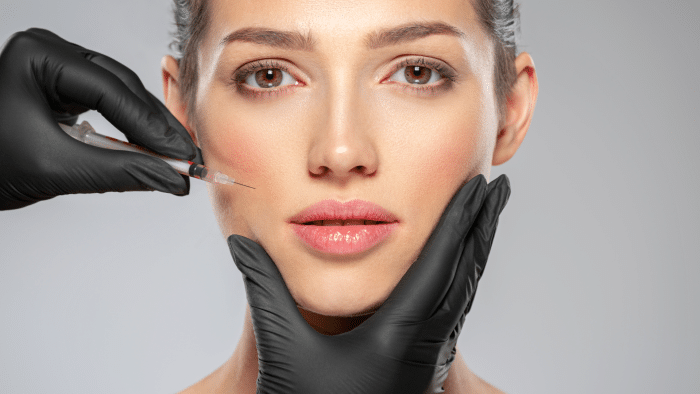 A person with a neutral expression receiving a Botox injection. Hands wearing black gloves are holding their chin and administering the injection with a syringe near the cheekbone. The background is a plain gray color, emphasizing the clinical environment and highlighting what not to do before such procedures.