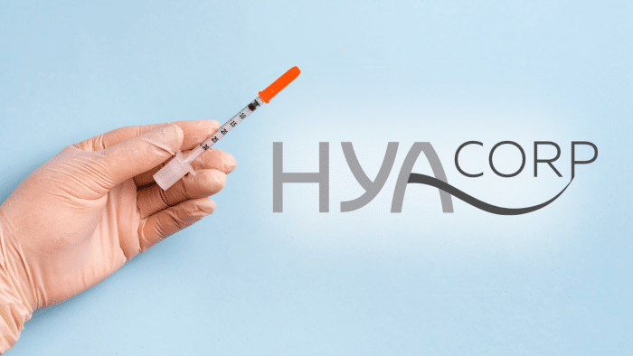 A gloved hand holds a syringe with an orange cap against a light blue background. On the right side of the image, the text "HYACORP" is displayed in a modern font, highlighting its prominence in hyacorp filler reviews.