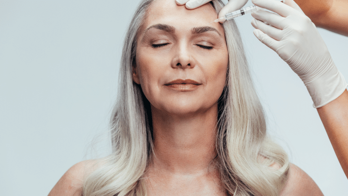 A woman with long, grey hair and closed eyes is receiving a cosmetic injection in her forehead from a practitioner wearing white gloves. Her expression appears calm and relaxed, leaving one to wonder at what age should Botox start to maintain such serene beauty.