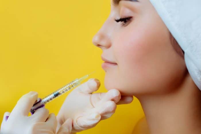 A woman with closed eyes and a towel wrapped around her head receives a cosmetic injection in her chin. A gloved hand holds her chin and another gloved hand holds a syringe filled with Dysport for smile lines. The background is a vibrant yellow.