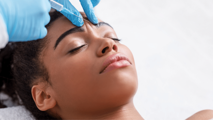 A close-up of a peaceful woman lying down with her eyes closed as a medical professional, indicated by their gloved hands, injects Juvederm into her forehead using a syringe. The background is blurred, focusing attention on the procedure.