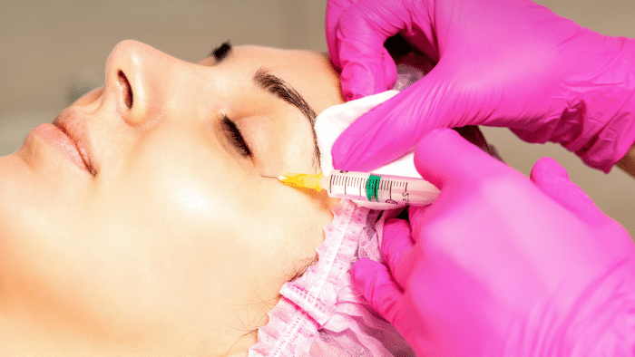 A person with eyes closed is receiving a Juvederm injection near the eye area to target under eye bags. The individual has a pink head covering and is being treated by hands in pink gloves holding a syringe. The procedure appears to be taking place in a clinical setting.