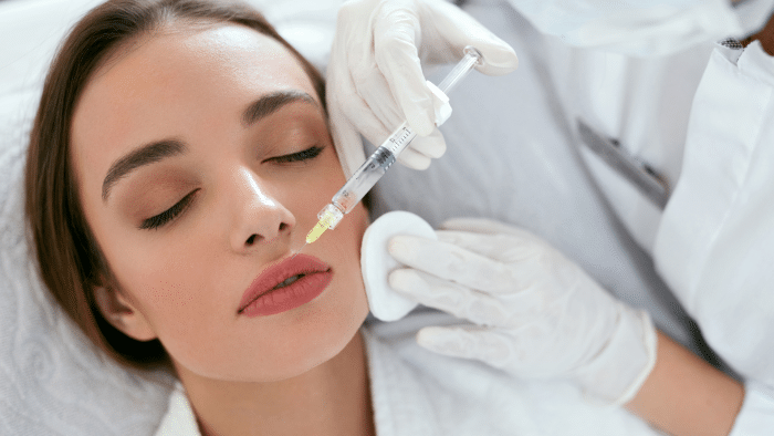 A woman with closed eyes is receiving a Restylane injection administered by a healthcare professional wearing white gloves. The professional holds a cotton pad on her face while injecting the substance with a syringe near her lips, ensuring minimal swelling.