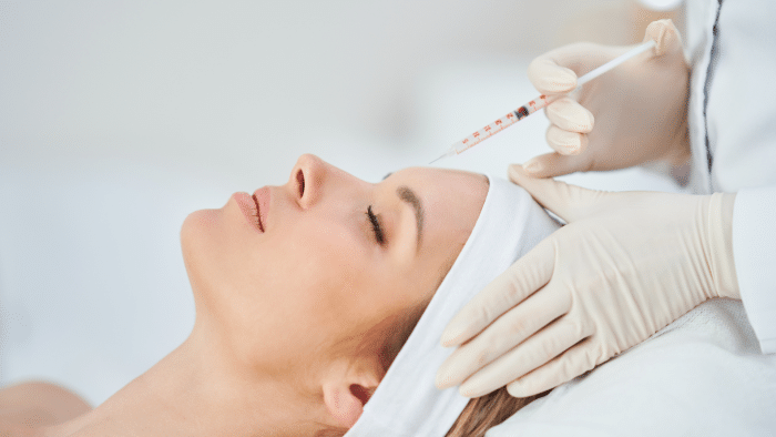 A person wearing a white headband and lying on their back receives an injection in their forehead from a professional in a white coat and latex gloves. The scene suggests a medical or cosmetic procedure, possibly involving Restylane filler, with consideration of potential side effects.