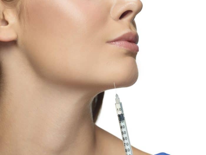 A close-up image of a woman's lower face and neck, with a syringe pointed at her chin. The woman has smooth, clear skin, reminiscent of patients receiving Dysport in the chin for a cosmetic or medical procedure. The background is white.