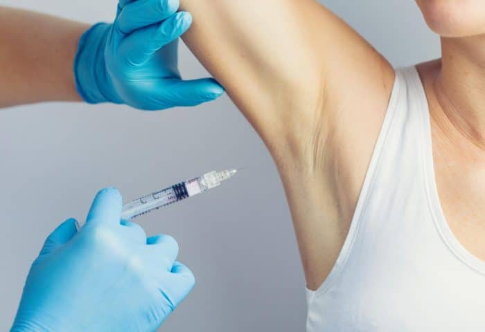 A person in a white tank top receives a Dysport injection for hyperhidrosis in the armpit area. The injector, wearing blue gloves, carefully holds the syringe with one hand and lifts the recipient's arm with the other.