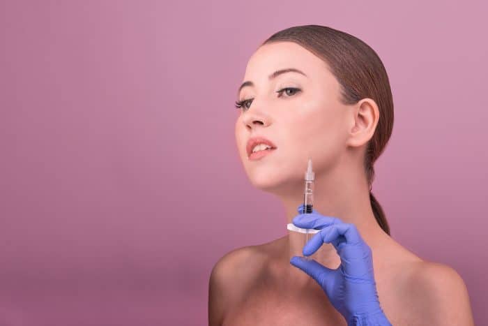 A woman with pulled-back hair stands against a pink background. She holds a syringe near her chin, looking upwards slightly. Wearing blue gloves, she appears to be preparing for a cosmetic procedure, possibly Dysport in masseter treatment.