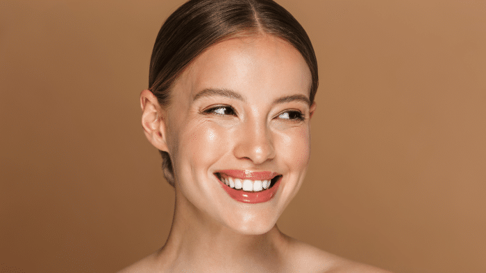 A person with smooth, glowing skin is smiling warmly. Their hair is slicked back neatly, and the background is a solid beige color. The lighting accentuates their radiant complexion, much like the results you can achieve with Restylane Contour vs Lyft treatments.
