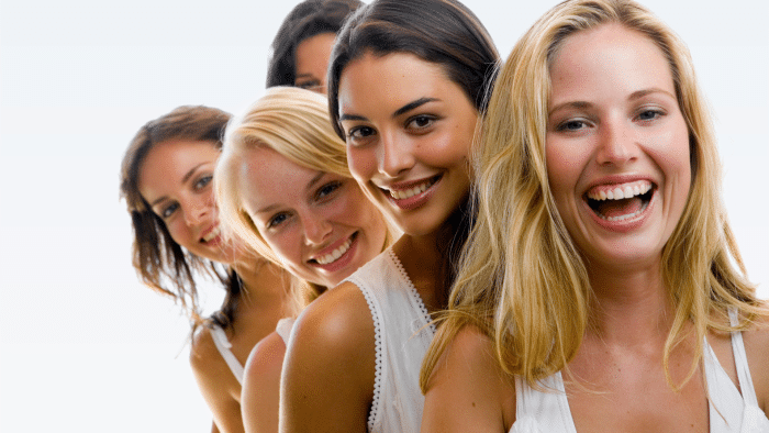 A row of five women standing closely together, each wearing white tops. They are all smiling and showing various expressions of happiness and excitement, possibly thanks to Restylane smoothing out their smile lines. The background is plain white, emphasizing the subjects.
