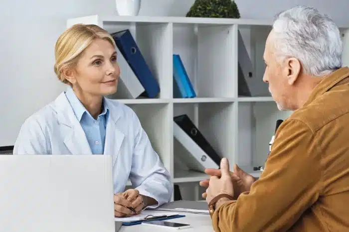 A female doctor in a white lab coat attentively listens to an older male patient seated in front of her. They are in a medical office with white shelves holding binders and a small potted plant in the background. The doctor has a laptop and clipboard on her desk.