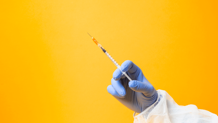 A hand in a blue glove, wearing a white medical gown, holds a syringe with an orange-tipped needle filled with Restylane ingredients against a bright orange background.