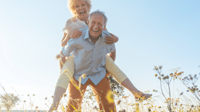 An elderly man is joyfully giving an elderly woman a piggyback ride in a sunlit field, both laughing and happy. Surrounded by tall grasses and wildflowers with clear blue skies in the background, their spirits seem as uplifted as if they'd just had an orthovisc injection for hip relief.