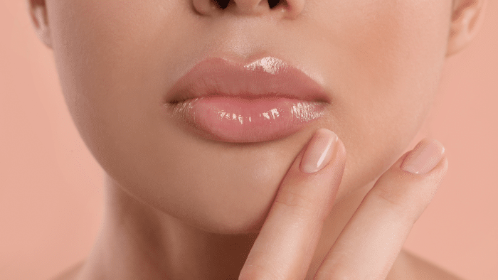 Close-up of a person’s face focusing on their full, glossy Restylane Silk lips. A hand with manicured nails lightly touches the chin. The background is a soft peach color, complementing the natural and elegant look.