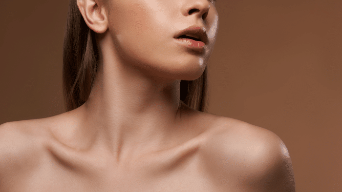 Close-up of the lower face and neck of a person with smooth skin, possibly enhanced by Restylane. The background is a solid brown color, emphasizing the well-defined jawline, lips, and collarbones.
