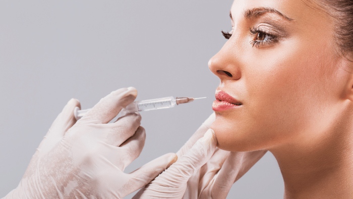 A close-up of a woman receiving a cosmetic injection on her lips. A medical professional wearing white gloves gently holds her chin and administers the injection with a syringe, possibly preparing for Restylane for jawline enhancement. The background is a plain, light grey color.