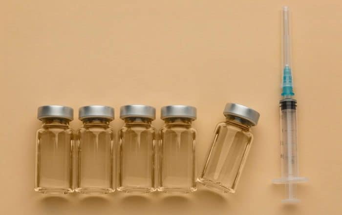 Five vials of clear liquid are lined up horizontally on a beige surface, with one vial leaning slightly to the right. Next to the vials is a syringe with a green-tipped needle, often used for Dysport treatments. What is it? Dysport helps reduce facial wrinkles and fine lines.