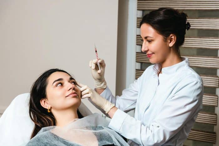 A woman in a medical setting is receiving an injection from a healthcare professional. The patient is lying back on a chair and looking up, while the healthcare professional, wearing gloves and a white coat, prepares the syringe with a focused expression, adhering closely to Dysport prescribing information.