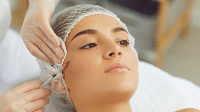 A person lies on a medical table, receiving an injection in the cheek. They wear a hair net, and the gloved hands of a professional are visible, holding the syringe filled with Restylane with lidocaine. The background is out of focus, suggesting a clinical setting.