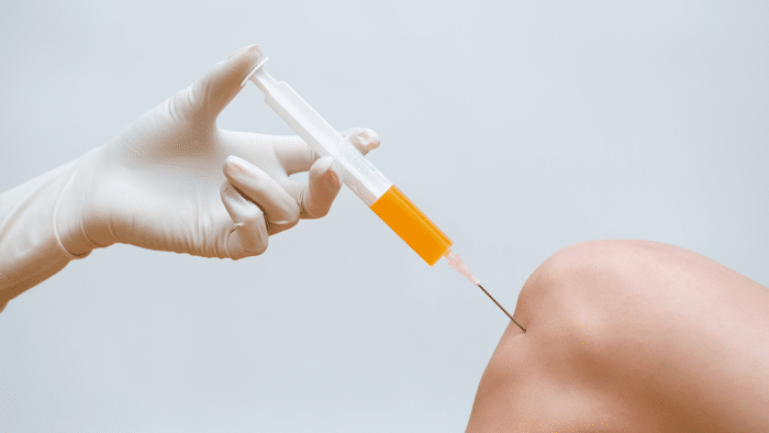 A gloved hand holds a syringe filled with yellow fluid, administering an injection into a person's knee against a light gray background, highlighting the ongoing debate of Orthovisc vs Synvisc for optimal treatment.