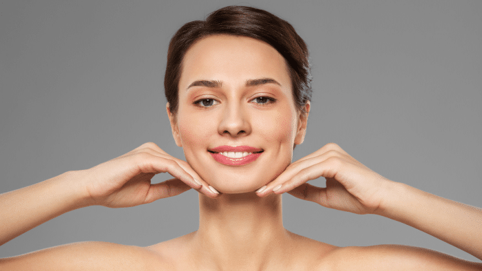 A person with short brown hair smiles and gently tilts their head upward, resting their chin on both hands. The background is a solid gray, and the person appears confident and relaxed, perhaps showing the results of a xeomin lip flip before and after.
