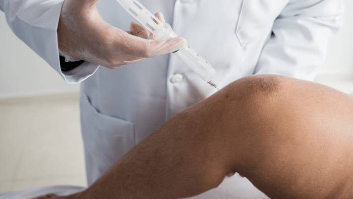 A healthcare professional in a white lab coat administers an injection to a patient's knee. The close-up image shows the syringe inserted into the knee area, suggesting a medical treatment or procedure such as Hyalgan vs Synvisc injections. The patient's leg is bent at the knee.