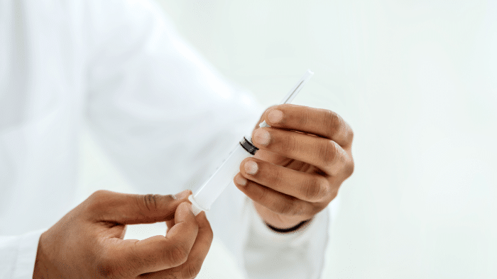A person wearing a white coat holds a syringe, preparing it for use, possibly for Juvederm. The background is blurred and white, giving a clean, clinical feel to the setting. The focus is on the hands and the syringe as they work meticulously to ensure minimal swelling during application.