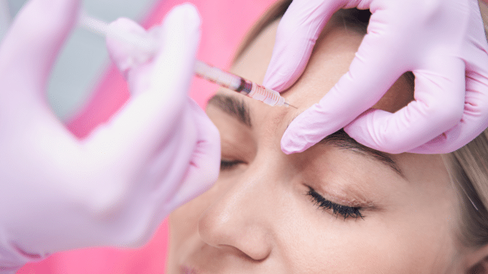 Close-up of a person receiving an injection in the forehead to treat Juvederm 11 lines. The person has their eyes closed and appears relaxed. The hands of the practitioner, wearing pink gloves, are gently holding the syringe and forehead. The background is blurred and pink.