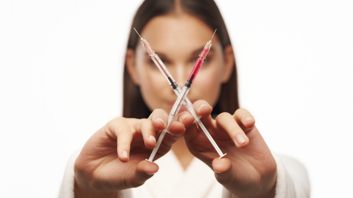 A person holds two syringes crossed in front of their face, with the needles pointing upwards. The syringes are filled with a red liquid, and the person's face is out of focus, emphasizing the syringes in the foreground—almost reminiscent of comparing xeomin vs botox which lasts longer. The background is plain white.