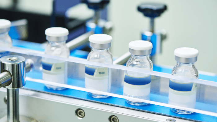 A close-up of multiple sealed vials with white stoppers on a blue production line conveyor belt in a laboratory. The clear vials contain a colorless liquid, and the entire setup appears to be part of a sterile manufacturing process at a leading xeomin manufacturer.