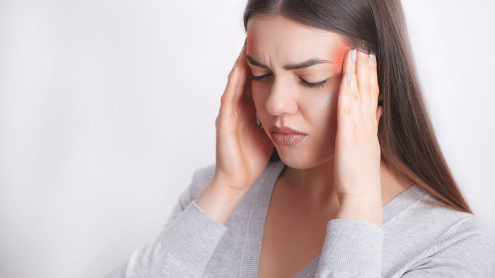 A woman with long dark hair holds her temples and has her eyes closed, appearing to be in pain or experiencing a headache, possibly in need of Xeomin for migraines. She is wearing a light-colored top against a plain white background.