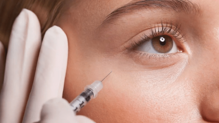 A close-up of a person's face focuses on their eye, with a gloved hand holding a syringe near the skin below, preparing for a Juvederm tear trough injection. The person has light skin, and the image captures the intricate details of their eye and eyelashes.