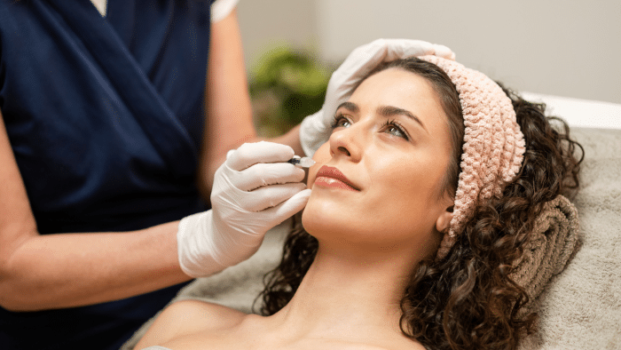 A woman with curly hair wearing a pink headband lies down with a towel behind her head. A person wearing gloves is holding a cosmetic tool close to her lips, preparing to apply makeup or identify xeomin injection sites. The woman looks relaxed and content.