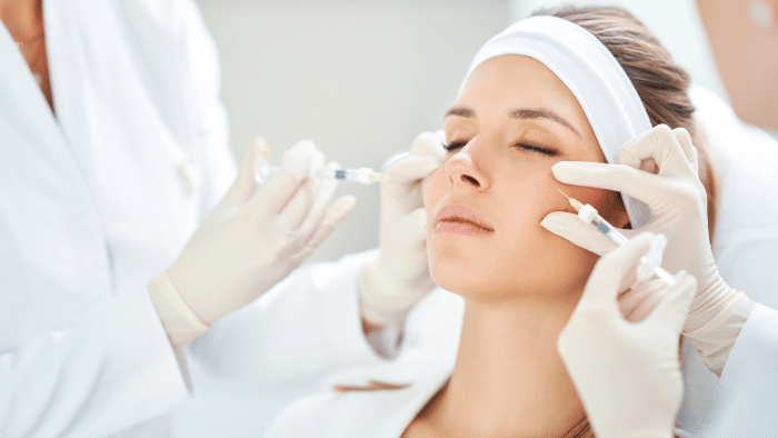 A woman with closed eyes and a relaxed expression receives Xeomin injections from medical professionals in white coats and gloves. A headband keeps her hair back as the procedure is performed on both sides of her face.