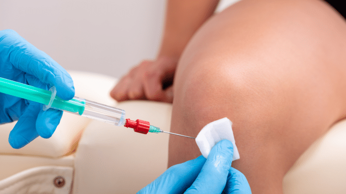 A healthcare professional wearing blue gloves is preparing to administer an Orthovisc knee injection. The professional is holding a syringe and using a cotton pad to clean the injection site on the patient's slightly bent knee, resting on a cushion.