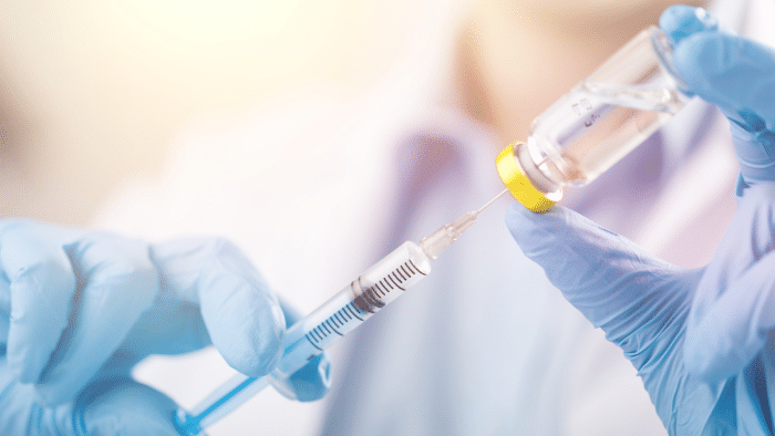 A person wearing blue gloves is holding a vial and drawing liquid from it into a syringe. The background is blurred, focusing on the hands performing the task, indicating a medical or vaccination process, possibly related to xeomin pros and cons.