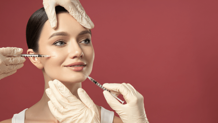 A woman with a serene smile is receiving Juvederm filler for jowls in both cheeks from two hands wearing white gloves against a solid red background. One hand is positioned on her forehead, steadying her head. She is wearing a white top.
