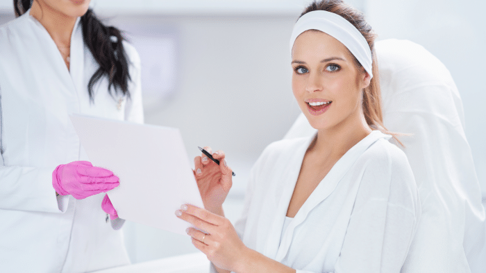 A woman in a white robe and headband holds a clipboard, smiling confidently at the camera. Beside her, another person in a white coat and pink gloves stands partially visible, suggesting a bright medical or spa setting where Juvederm vs Kysse options might be discussed.