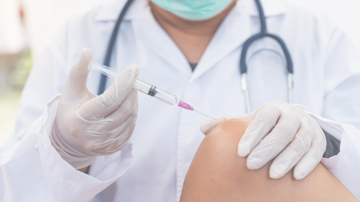 A healthcare professional wearing a white coat, gloves, and a stethoscope prepares to give an injection of Hyalgan to a patient's knee. The professional holds a syringe with pink liquid, positioned close to the patient's bare knee.