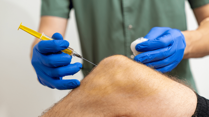 A medical professional wearing a green shirt and blue gloves is injecting a yellow fluid into a patient's knee using a syringe, likely as part of the Hyalgan vs Orthovisc treatment. The practitioner's other hand holds a piece of gauze next to the injection site. The focus is on the knee and hands.