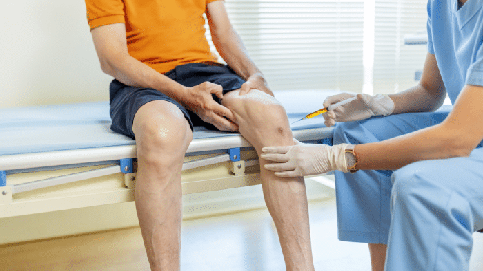 A healthcare professional in blue scrubs and gloves administers an injection to an elderly person’s knee, who sits on an examination table in an orange shirt and dark shorts. The setting appears to be a medical clinic, possibly discussing treatment options like Orthovisc vs Euflexxa for joint pain relief.
