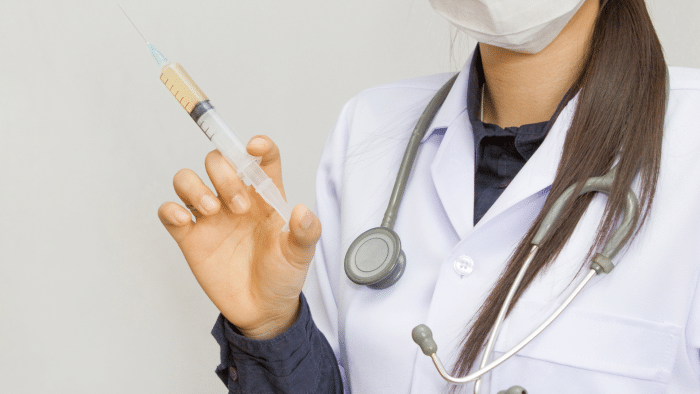 A healthcare professional wearing a white lab coat and stethoscope holds a syringe filled with liquid, likely prepared for a hyalgan dosage. The individual's face is partially obscured by a mask. The background is plain and light-colored.