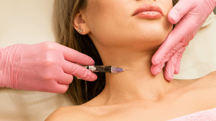 A person receiving an injection in the neck area administered by someone wearing pink gloves. The person has a calm expression with their eyes closed. The syringe contains a clear liquid, likely Juvederm for neck treatment. The background is neutral, focusing on the procedure.