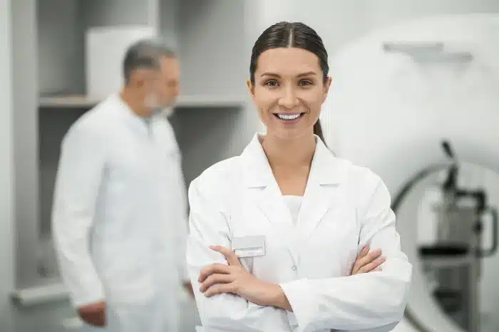 A woman in a white lab coat smiles confidently with her arms crossed, standing in a medical or laboratory setting. Behind her, a man in a similar white lab coat is slightly blurred and out of focus. Medical equipment is visible in the background.