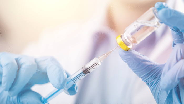 Close-up of a person wearing blue gloves holding a syringe and drawing a clear liquid from a vial with a yellow cap. The background is blurred, highlighting the hands and medical equipment, possibly used for Juvederm treatments, in focus. Always consult your doctor regarding any allergy concerns.