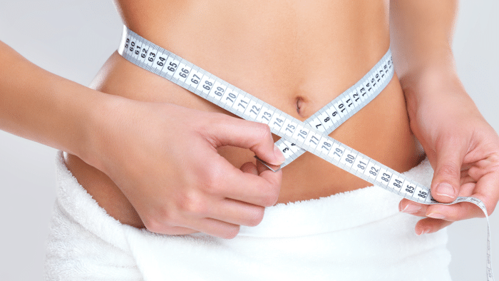 A person dressed in a white towel measures their waist with a white measuring tape. The tape is wrapped around their bare midsection, displaying measurements in centimeters. Their fingers are adjusting the tape to get an accurate reading.