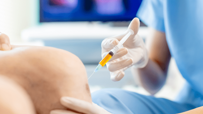 A healthcare professional, wearing blue scrubs and white gloves, prepares to administer an injection into the arm of a patient. The focus is on the syringe and the point of contact on the patient's arm. Background details are blurred.