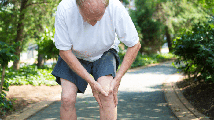 An elderly man in a white shirt and gray shorts is bending over, holding his knee with both hands, appearing to be in discomfort or pain. He is standing on a pathway surrounded by greenery and trees, with the path stretching into the distance.