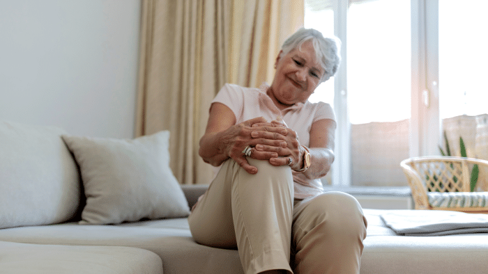 A senior woman with short white hair is sitting on a couch and holding her knee with both hands. She appears to be in pain. She is wearing a light-colored shirt and beige pants. The room is bright with a large window and light-colored curtains in the background.