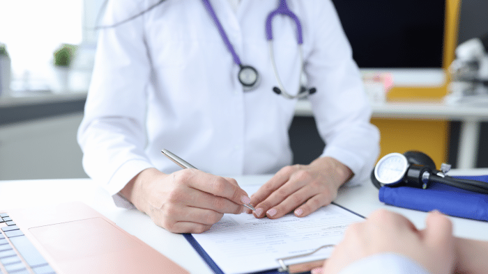 A medical professional wearing a white coat and stethoscope around the neck is writing on a clipboard. The person's hands are visible, and another individual's hands are seen across the table. A laptop and medical equipment are on the desk.